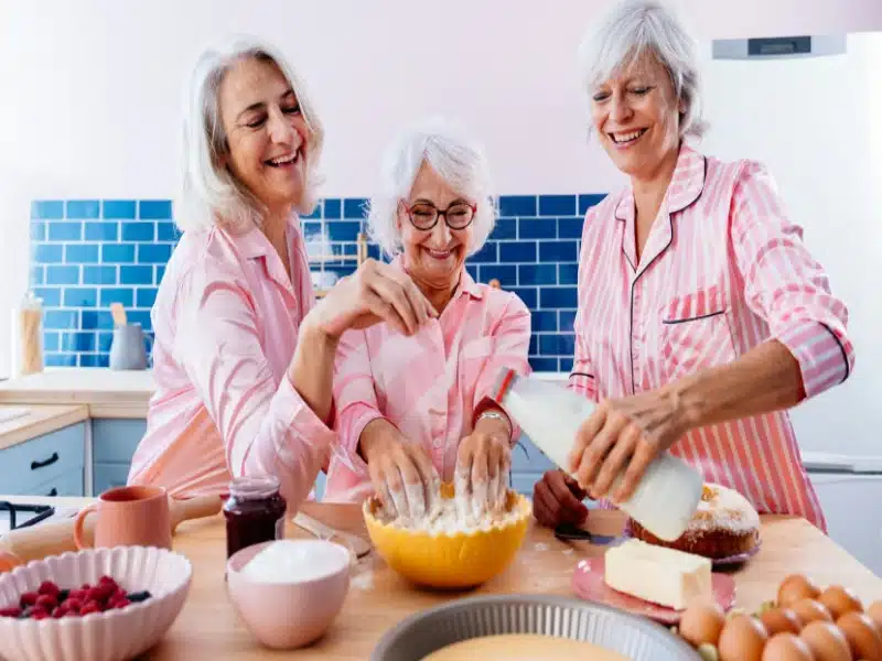 Three elderly women in a home kitchen, smiling while preparing food together, wearing matching pink striped pajamas.